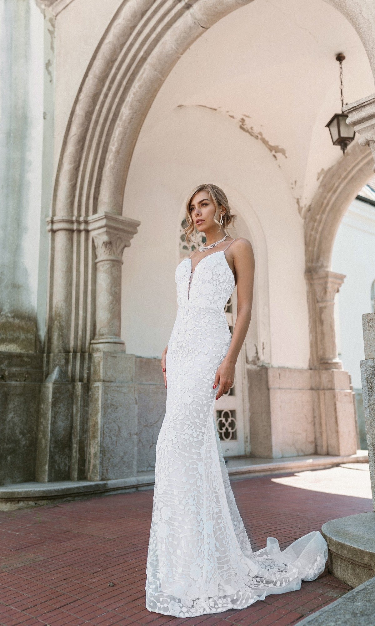 A woman wearing the Tina Holly BB008 Long White Lace Formal Dress stands outdoors before an arched stone structure, gazing slightly to the side.